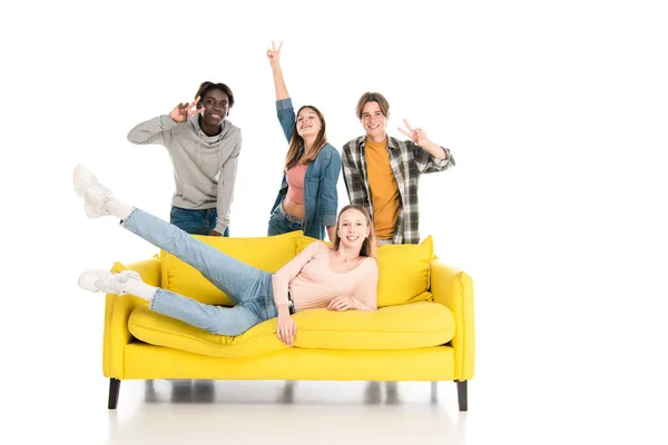 Adolescentes alegres multiétnicos mostrando gesto de paz no sofá amarelo no fundo branco — Fotografia de Stock