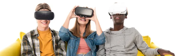Panoramic shot of smiling multiethnic teenagers using vr headsets on couch isolated on white — Stock Photo