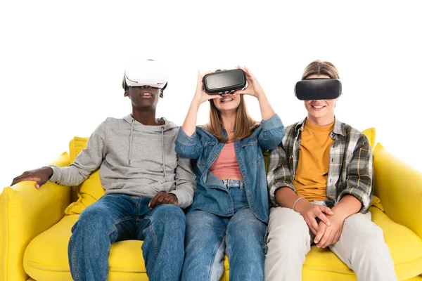 Smiling multicultural teenagers in vr headsets sitting on yellow couch isolated on white — Stock Photo