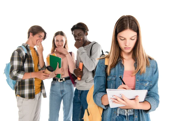 Enfoque selectivo de la niña escribiendo en un cuaderno cerca de adolescentes multiétnicos sonrientes aislados en blanco - foto de stock