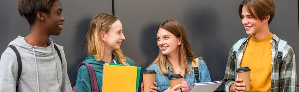 Panoramic shot of smiling multiethnic teenagers with notebooks and coffee to go standing near building — Stock Photo