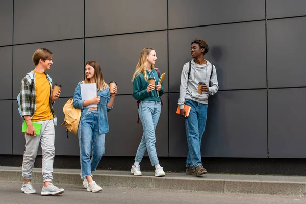 Adolescentes multiétnicos sorridentes com café para ir e cadernos andando perto do prédio ao ar livre — Fotografia de Stock