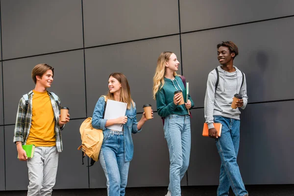 Positive multiethnic teenagers with backpacks and coffee to go walking near building — Stock Photo