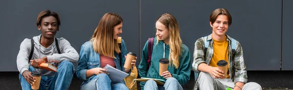 Panoramic shot of multiethnic teenagers with coffee to go and notebooks talking near building — Stock Photo