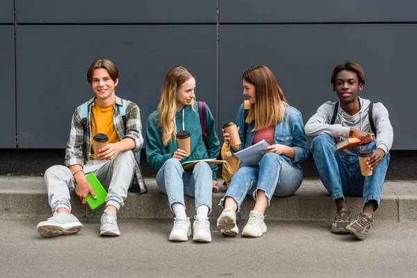 Sonrientes adolescentes multiétnicos con café para llevar y mochilas hablando cerca del edificio - foto de stock