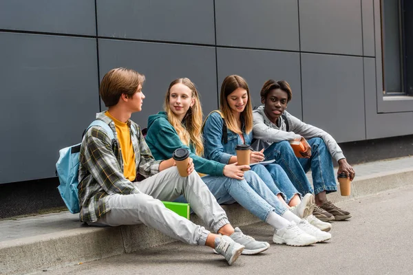 Positive multiethnic teenagers with coffee to go and books sitting on sidewalk near building — Stock Photo