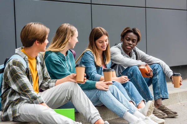 Focus selettivo di sorridente adolescente multietnico con caffè per andare e libri seduti vicino a un amico che scrive su notebook e edificio — Foto stock