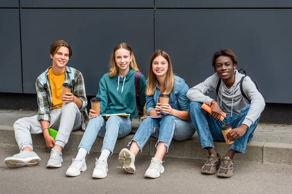 Cheerful multiethnic teenagers with coffee to go and notebooks looking at camera on sidewalk near building — Stock Photo