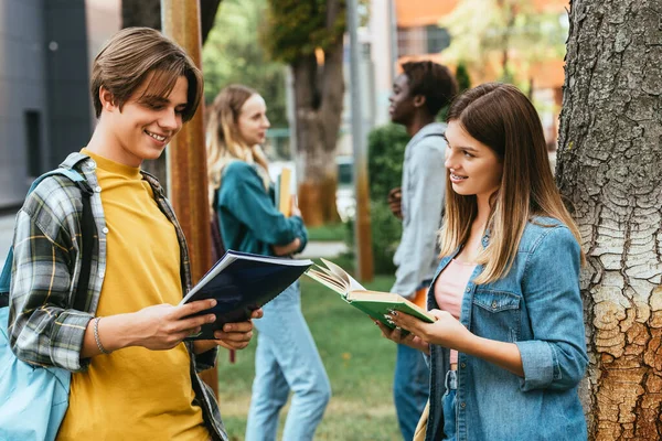 Foco seletivo de adolescentes sorridentes com livro e caderno em pé perto da árvore ao ar livre — Fotografia de Stock