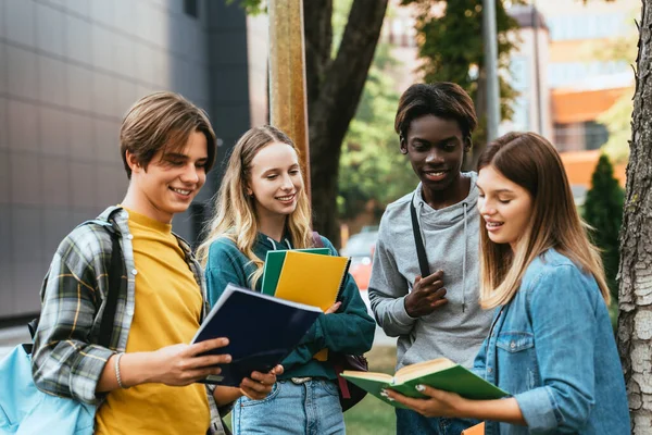 Selective focus of positive multiethnic teenagers looking at book near tree outdoors — Stock Photo