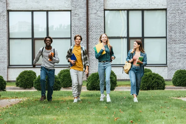 Selective focus of cheerful multiethnic teenagers with notebooks and backpacks walking on grass outdoors — Stock Photo