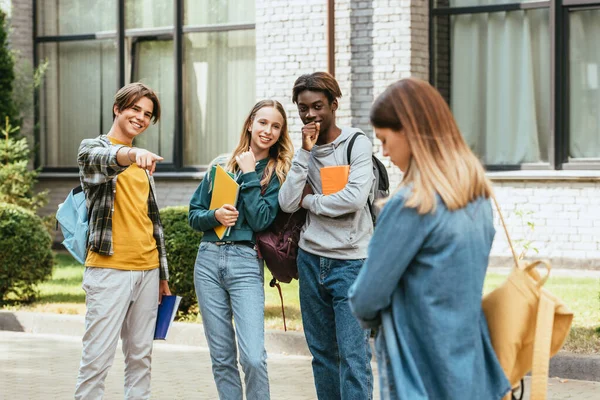 Selective focus of smiling multicultural teenagers with backpacks pointing at sad girl outdoors — Stock Photo