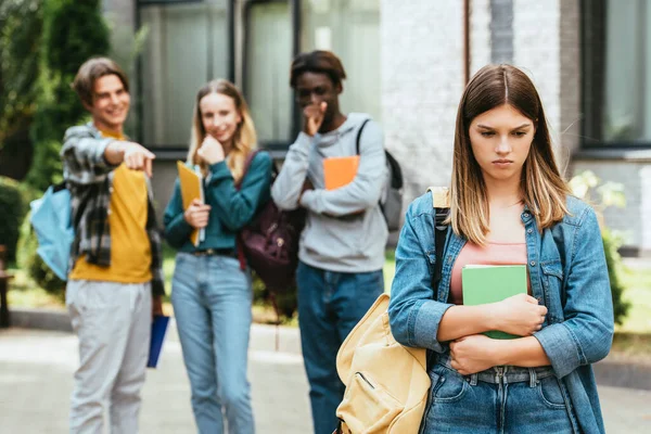 Selective focus of sad teenager with book and backpack standing near multiethnic schoolkids pointing with finger outdoors — Stock Photo