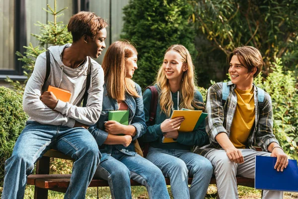 Sonrientes adolescentes multiculturales con libros hablando en el banquillo al aire libre - foto de stock