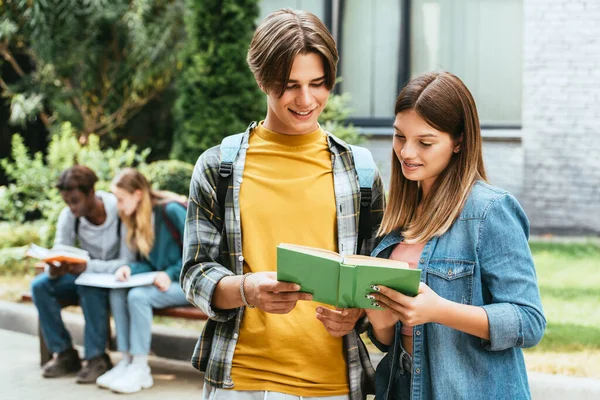 Selective focus of smiling teenagers reading book outdoors — Stock Photo