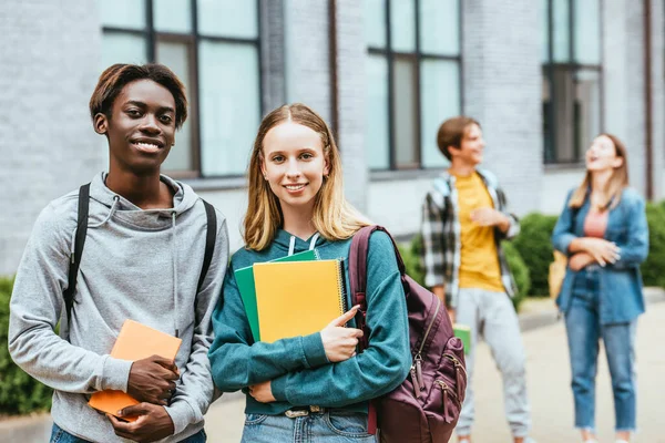 Selektiver Fokus lächelnder multiethnischer Teenager mit Rucksäcken und Notizbüchern, die im Freien in die Kamera schauen — Stockfoto