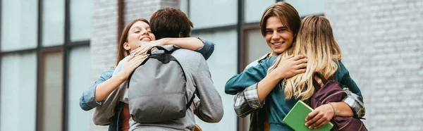 Orientación panorámica de adolescentes sonrientes multiculturales con mochilas abrazándose al aire libre - foto de stock