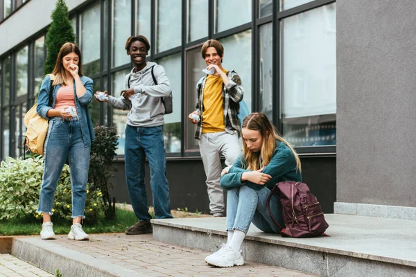 Enfoque selectivo de chica triste con mochila cerca de adolescentes multiétnicos sonrientes con papel agrupado al aire libre - foto de stock