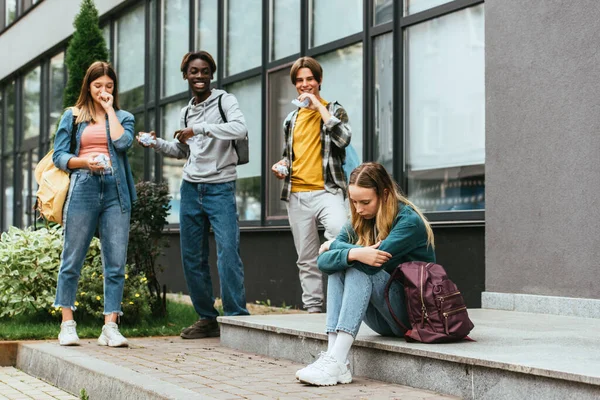 Enfoque selectivo de chica triste con mochila cerca de adolescentes multiétnicos sonrientes con papel agrupado al aire libre — Stock Photo