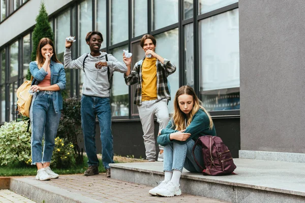 Enfoque selectivo de chica molesta cerca de adolescentes multiculturales sonrientes con papel agrupado al aire libre - foto de stock