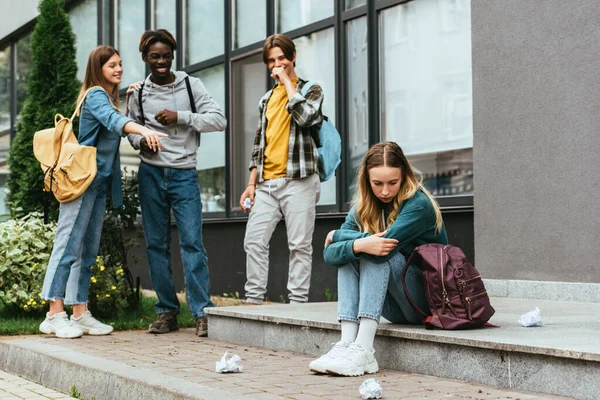 Foco seletivo da menina triste sentada perto da mochila, papel amarrotado e adolescentes multiétnicos sorridentes ao ar livre — Fotografia de Stock