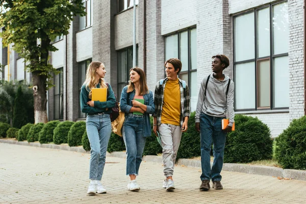 Smiling multicultural teenagers with notebooks and backpacks walking on urban street — Stock Photo