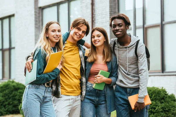 Adolescents multiethniques souriants avec des livres embrassant à l'extérieur — Photo de stock