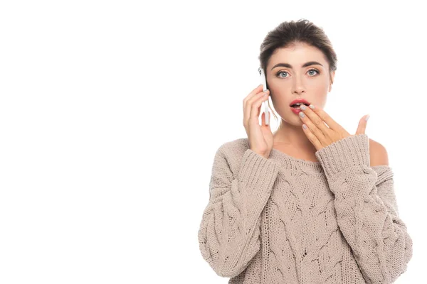 Schockierte Frau im eleganten durchbrochenen Pullover, der Mund mit der Hand verdeckt, während sie auf dem Smartphone isoliert auf weiß spricht — Stockfoto