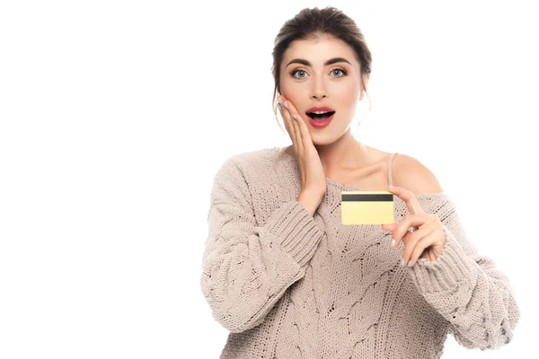 Mujer conmocionada en suéter de punto tocando la cara mientras sostiene la tarjeta de crédito aislada en blanco - foto de stock