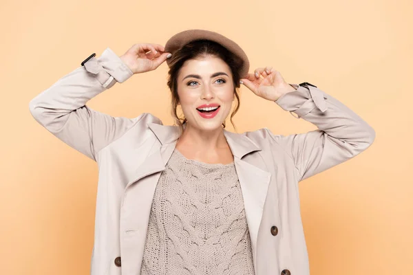 Stylish woman in trench coat touching beret while looking away on peach — Stock Photo