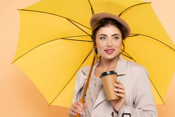 Веселая женщина в плаще и берете держит одноразовую чашку под желтым зонтиком на персике — стоковое фото