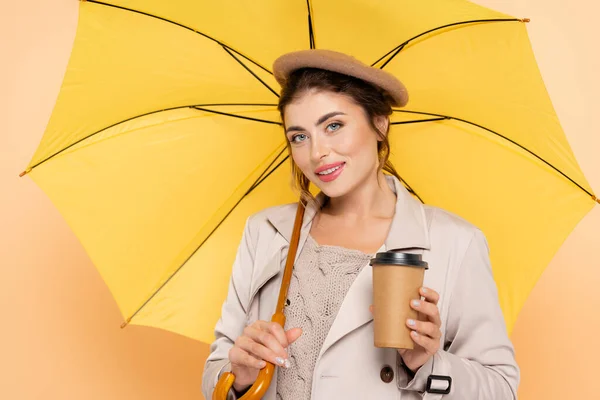 Femme à la mode en trench coat et béret tenant café pour aller sous parapluie jaune sur la pêche — Photo de stock