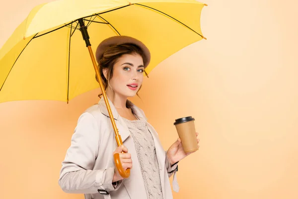 Sensual woman in stylish autumn outfit looking at camera while holding coffee to go under yellow umbrella on peach — Stock Photo