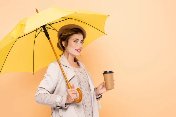 Elegant woman in trench coat and beret holding paper cup under yellow umbrella on peach — Stock Photo