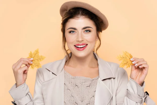 Fashionable woman in autumn outfit holding yellow leaves while looking at camera isolated on peach — Stock Photo