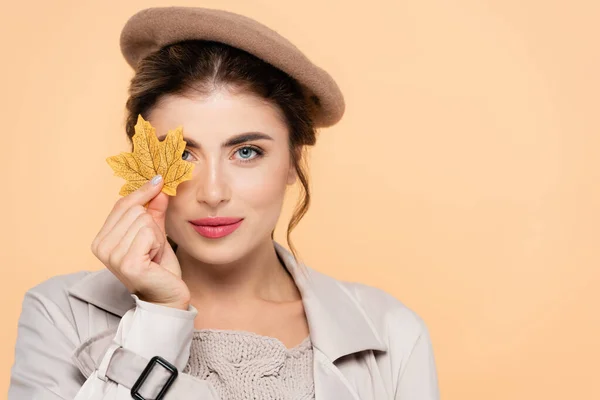 Модная женщина в осеннем наряде закрывает глаза желтым листом, глядя на камеру, изолированную от персика — стоковое фото