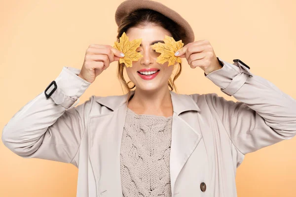 Joyeuse femme en élégant trench coat et béret couvrant les yeux avec des feuilles jaunes isolées sur la pêche — Photo de stock