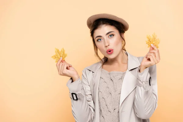 Здивована жінка в модному осінньому вбранні, що тримає жовте листя ізольовано на персику — стокове фото
