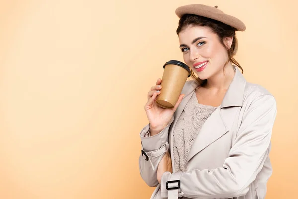 Mujer joven en gabardina y boina sosteniendo café para llevar mientras mira la cámara aislada en melocotón - foto de stock
