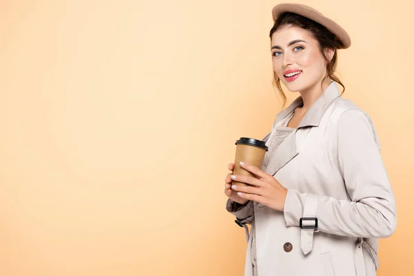 Mujer de moda en traje de otoño sosteniendo café para ir mientras mira la cámara aislada en melocotón - foto de stock