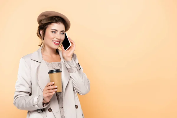 Mujer joven en traje de otoño elegante celebración de café para llevar mientras habla en el teléfono inteligente aislado en melocotón - foto de stock