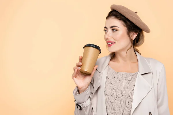 Mujer de moda en traje de otoño sosteniendo café para ir mientras mira hacia otro lado aislado en melocotón - foto de stock