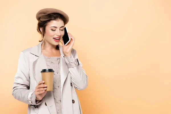 Trendy woman in autumn outfit talking on mobile phone while holding coffee to go isolated on peach — Stock Photo