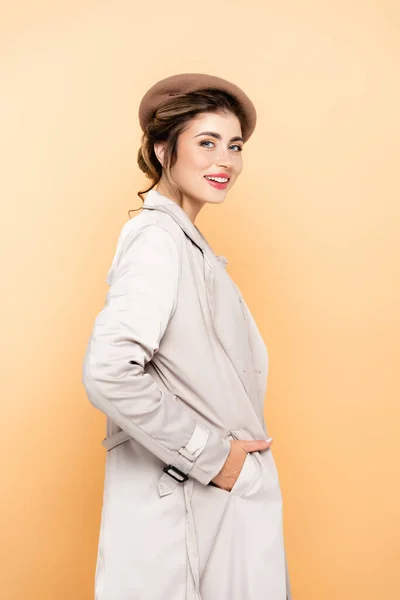 Trendy woman in beret and trench coat looking at camera while posing with hand in pocket on peach — Stock Photo