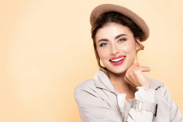 Joyful, fashionable woman in beret holding hand near chin while looking at camera  isolated on peach — Stock Photo