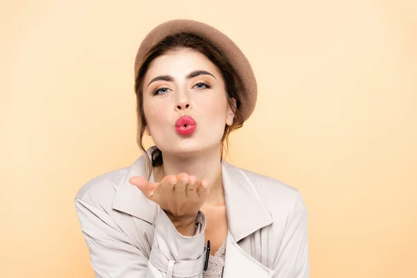 Sensual woman in trench coat and beret blowing air kiss at camera isolated on peach — Stock Photo