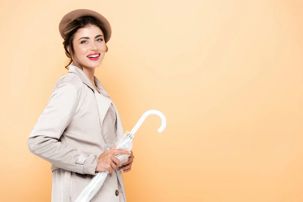 Joyful woman in stylish autumn outfit holding folded umbrella while looking at camera on peach — Stock Photo