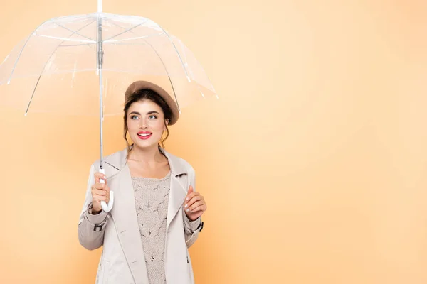 Fröhliche Frau in Trenchcoat und Baskenmütze, die unter einem durchsichtigen Regenschirm auf Pfirsich wegschaut — Stockfoto