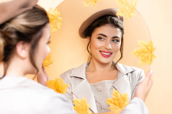 Enfoque selectivo de la mujer joven en gabardina y boina mirando en el espejo decorado con hojas de otoño en melocotón - foto de stock