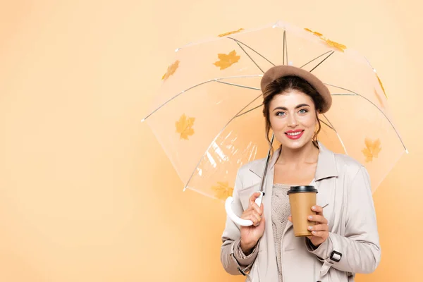 Mujer alegre en traje de otoño elegante celebración de café para ir bajo paraguas con follaje en melocotón - foto de stock